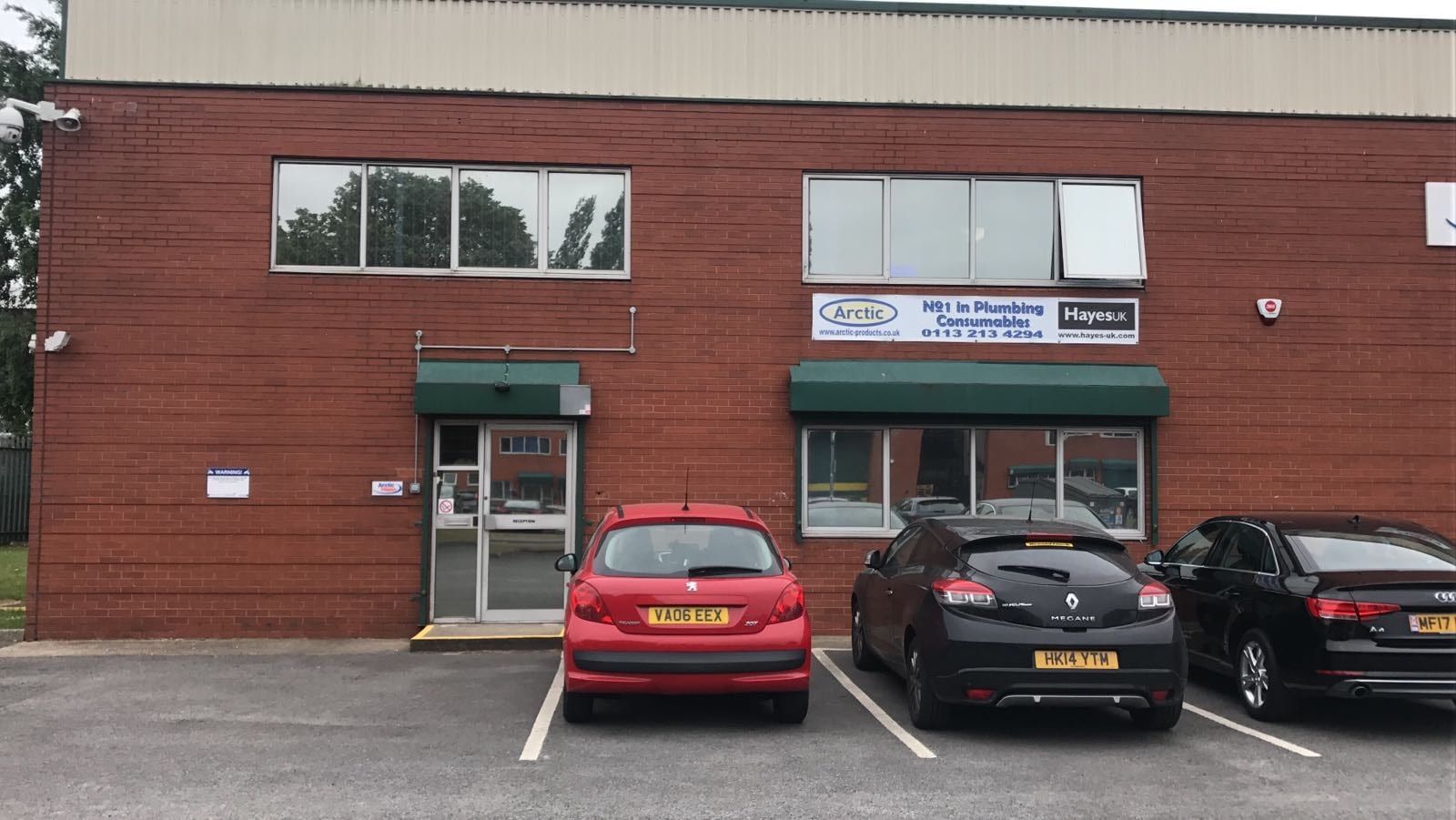 Red-brick commercial unit with green window awnings and an 'Arctic — Hayes UK' plumbing-consumables sign in the upper window, cars parked outside.