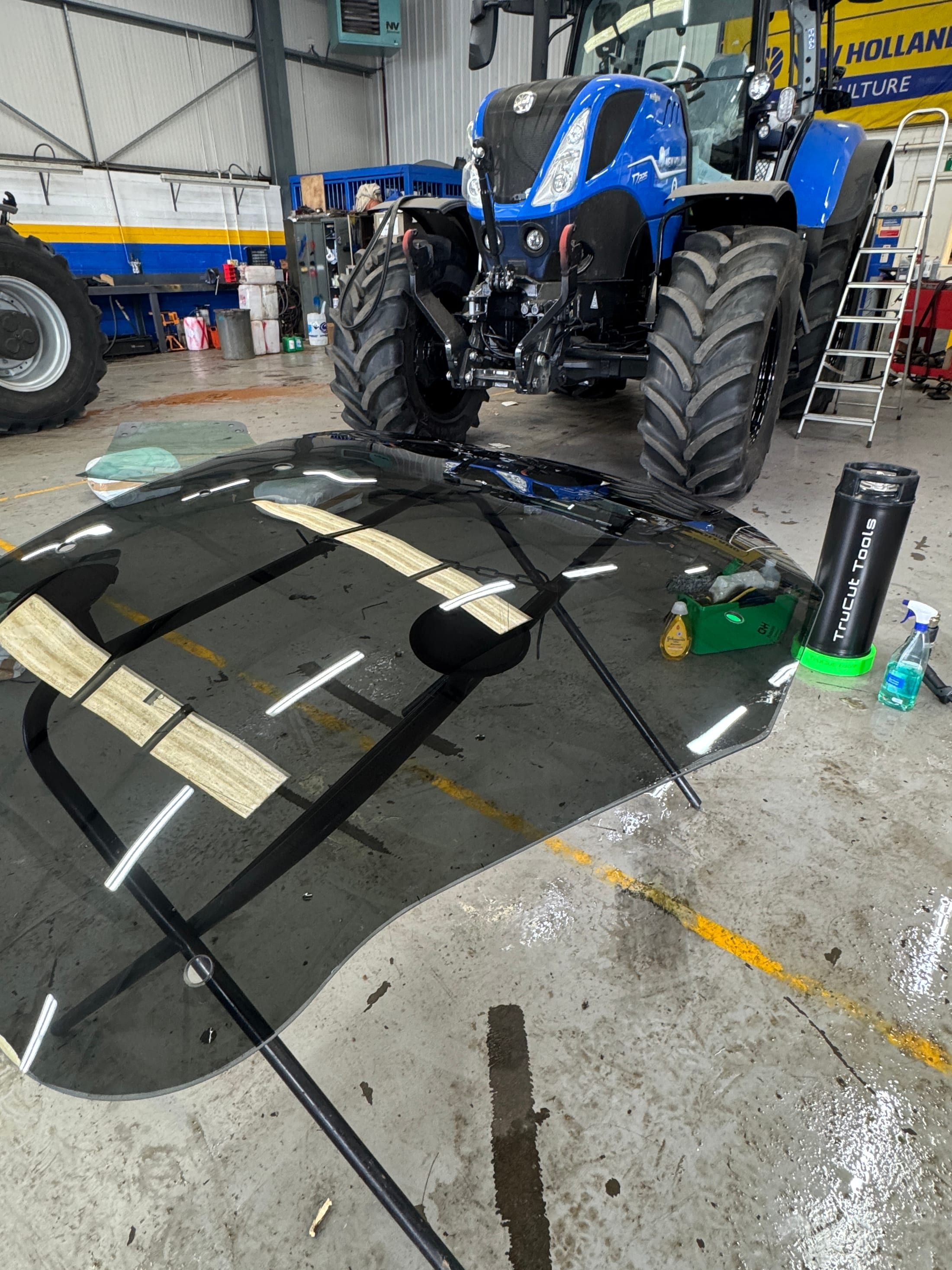 Tractor windscreen laid flat on a workshop floor mid-tint application, a blue New Holland tractor visible in the background.